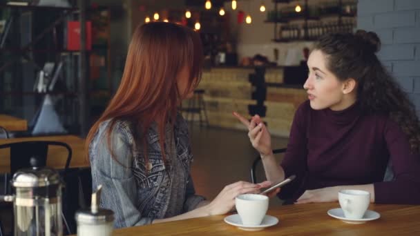 Young women are using smartphone, talking and doing high five while drinking tea in cafe in lunchtime. Communication, modern technology, girl time and happy people concept. — Stock Video