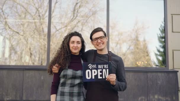 Portret van vrienden zakenpartners openen restaurant ingedrukt te houden "we zijn open" teken voor raam buiten het gebouw. Succesvolle start-up concept. — Stockvideo