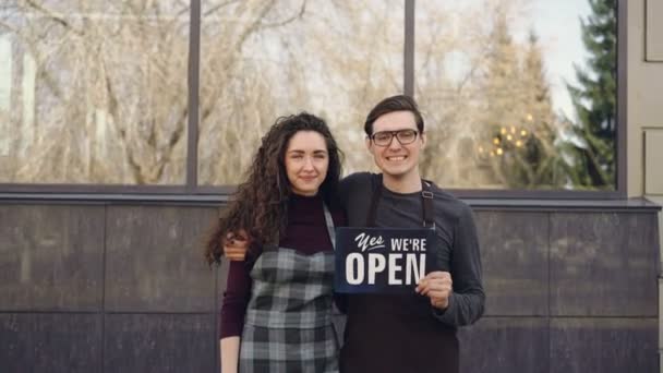 Retrato de parejas casadas felices socios de negocios abriendo café y sosteniendo "estamos abiertos" tablero en frente de la ventana exterior del edificio. Concepto de puesta en marcha exitoso . — Vídeos de Stock