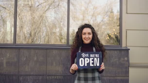 Portret van vrolijke mooie serveerster in schort holding "Ja we zijn open" teken permanent buiten en glimlachend camera kijken. Vanaf catering bedrijfsconcept. — Stockvideo