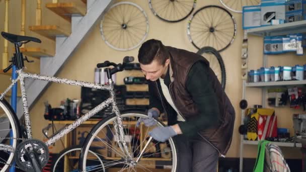 Knappe mannelijke mechanic is repareren wiel van de fiets met professioneel hulpmiddel terwijl werkt alleen in kleine werkplaats met scheepsuitrusting en reserveonderdelen zichtbaar. — Stockvideo