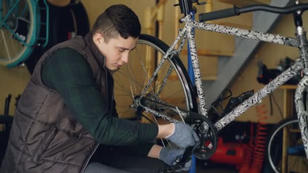 Jeune homme qualifié mécanicien professionnel fixe la bande de roulement de vélo avec la clé assis près du vélo sur le sol de son lieu de travail. Maintenance, outils et concept humain . — Video