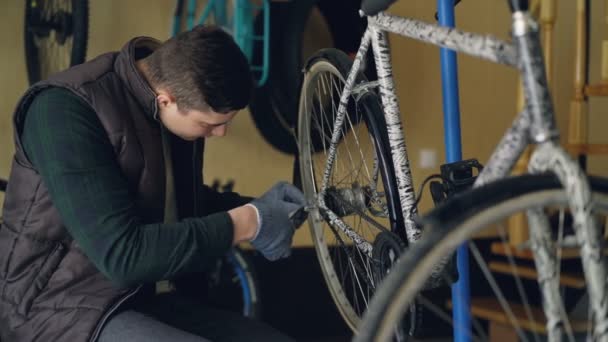 Handsome male serviceman in gloves is repairing back wheel of bicycle using professional tools. Bike spare parts and small workshop are visible. — Stock Video