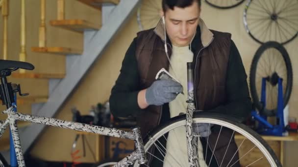 Le jeune mécanicien concentré grasse la roue du vélo et écoute de la musique avec des écouteurs tout en réparant le vélo dans son petit studio. Maintenance et concept de personnes . — Video