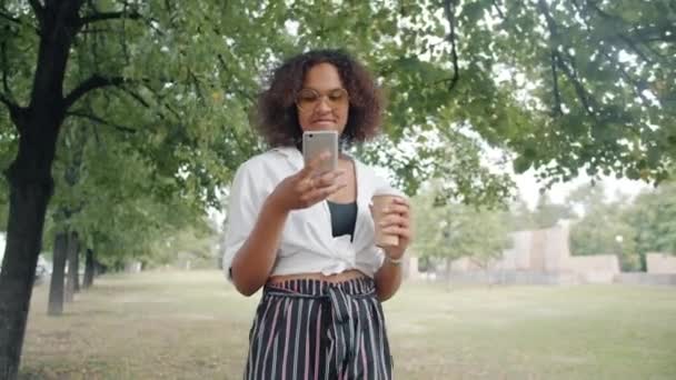 非洲裔美国女士在城市公园用智能手机边走边喝咖啡 — 图库视频影像