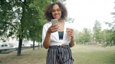 Afro-Amerikalı kadın kahve içiyor ve parkta tek başına akıllı telefon kullanıyor.