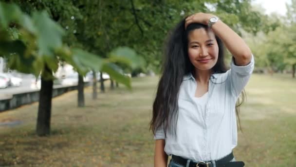 Портрет привлекательной азиатки, улыбающейся трогательными волосами на открытом воздухе в городском парке — стоковое видео