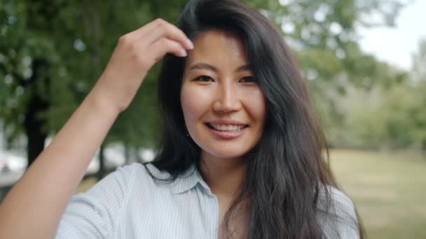 Захопливий портрет азіатської жінки, яка дивиться на камеру посміхаючись зачіски в парку — стокове відео