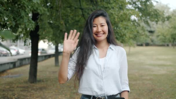 Медленный портрет азиатки, машущей рукой, улыбающейся на открытом воздухе в городском парке — стоковое видео
