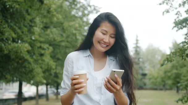 Χαριτωμένο Ασιάτης φοιτητής περπάτημα στο πάρκο με καφέ και smartphone πόσιμο — Αρχείο Βίντεο