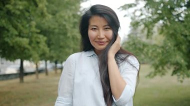 Neşeli Asyalı öğrenci parkta flört ediyor saçlarına dokunuyor kameraya bakıp gülümsüyor.