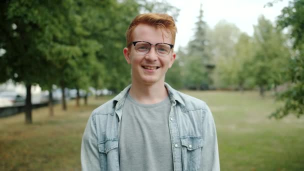 Slow motion of handsome redhead guy in glasses smiling in city park in summer — Stockvideo