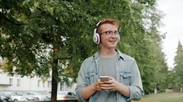 Kulaklıklı mutlu adam parkta yürüyüp akıllı telefon kullanarak müzik dinliyor. — Stok video