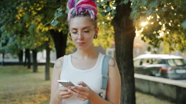 Şehir parkında akıllı telefon kullanarak gülümseyen, renkli saçlı tatlı bir kız. — Stok video