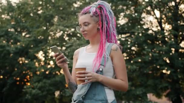 Jolie étudiante avec des cheveux colorés en utilisant smartphone tenant café à l'extérieur — Video