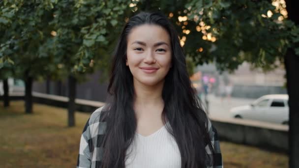 Güzel Asyalı öğrenci şehir parkında el hareketi yaparken gülümsüyor.