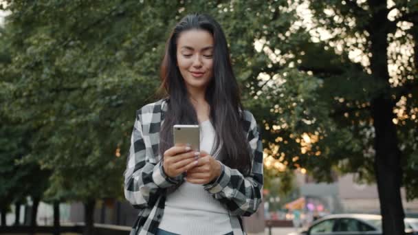 Şehir parkında akıllı telefon ekranına dokunan tatlı Asyalı kız tek başına gülümsüyor. — Stok video