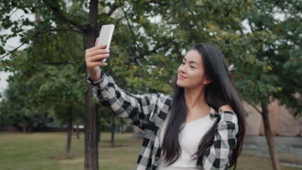 Портрет привлекательной азиатки, делающей селфи в парке с камерой на смартфоне — стоковое видео