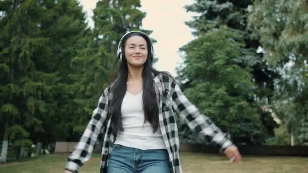 Счастливый азиатский студент танцует на открытом воздухе в беспроводных наушниках улыбаясь — стоковое видео