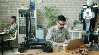 Ofiste dizüstü bilgisayarı kullanan genç geliştirici, 3D yazıcı ve robot görünür