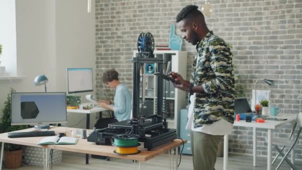 Νεαρός που χρησιμοποιεί την εκτύπωση tablet 3d μέρος για ρομπότ που εργάζονται στο γραφείο απολαμβάνοντας την εργασία — Αρχείο Βίντεο
