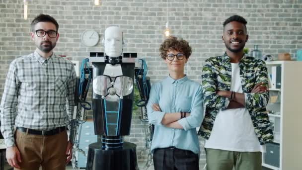 Retrato de ingenieros sonrientes y robot mirando a la cámara de pie en la oficina — Vídeos de Stock