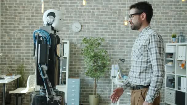 Молодой мужчина-разработчик и мужественный робот-киборг пожимает руку стоя на своем посту — стоковое видео