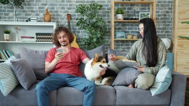 Mutlu genç çift kanepede köpekle dinleniyor, adam akıllı telefon kullanıyor, kız okuyor. — Stok video