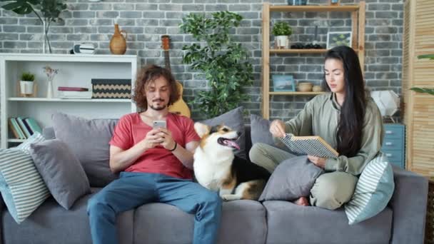 Чоловік використовує смартфон, коли жінка грабує собаку і читає книгу в квартирі — стокове відео