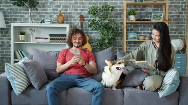 Szczęśliwa para korzystających dzień w domu za pomocą smartfona czytania książki głaskanie psa — Wideo stockowe