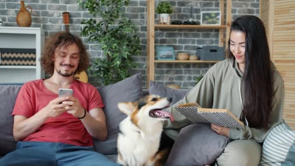 Guy bemutató okostelefon képernyőn a lány olvasás könyv simogató kutya otthon — Stock videók