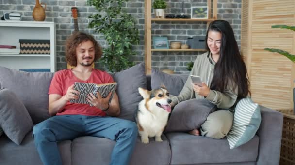 Mädchen mit Smartphone, während Kerl zu Hause auf Couch mit Corgi-Welpe Buch liest — Stockvideo