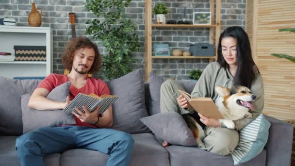 Jovem marido e mulher lendo livros em casa e acariciando filhote de cachorro corgi — Vídeo de Stock