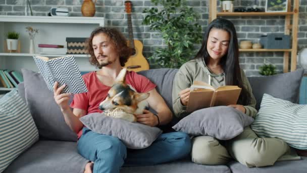 Fille et gars lisant des livres tandis que le chiot corgi mâche jouet sur le canapé à la maison — Video