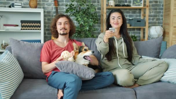 Alegre pareja viendo televisión en casa y acariciando corgi perrito en apartamento — Vídeos de Stock