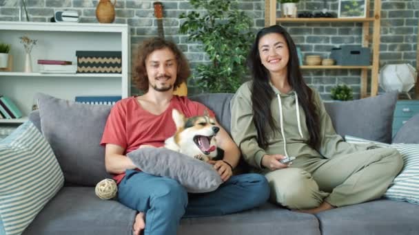 Multi-ethnisches Paar sieht Film im Fernsehen lächelnd Corgi Hündchen zu Hause streicheln — Stockvideo
