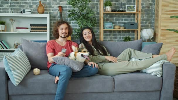 Hombre y mujer viendo comedia en la televisión riendo en el sofá en casa con lindo perro — Vídeos de Stock