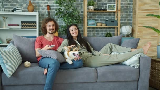 Mladá rodina a corgi pes sledování televize na gauči doma smát relaxaci — Stock video