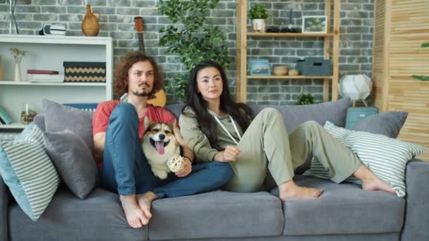 Asiático chica y caucásico chico viendo televisión en casa jugando con corgi perro — Vídeos de Stock