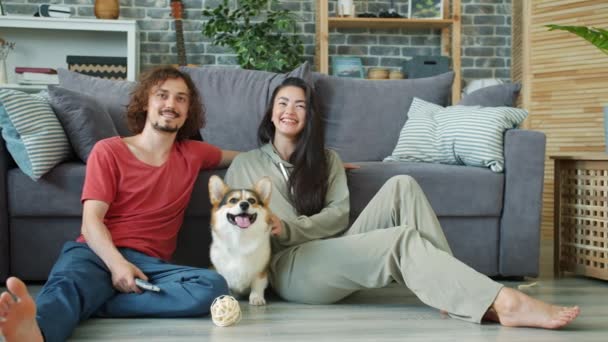 Φιλενάδα, φίλος και σκύλος βλέποντας τηλεόραση κάθεται στο πάτωμα στο διαμέρισμα χαμογελώντας — Αρχείο Βίντεο