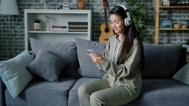 Genç bir kadın kulaklıkla müzik dinliyor ve evde akıllı telefon kullanıyor.