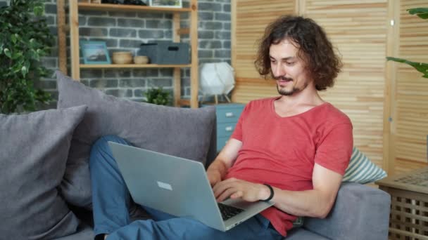 Chico sonriente usando el ordenador portátil en el sofá en casa escribiendo sonriendo trabajando en línea — Vídeos de Stock