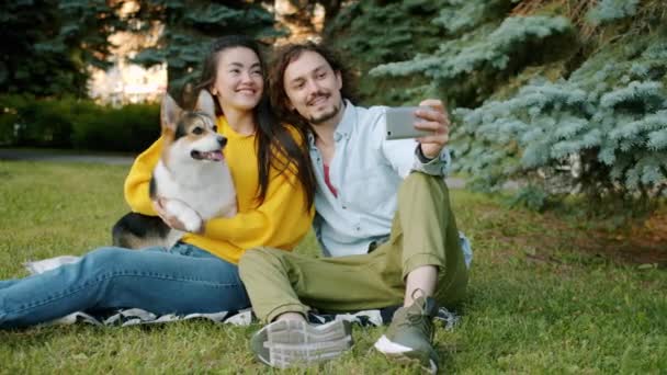 Ευτυχισμένο ζευγάρι λήψη selfie με corgi σκυλί φυλής στο πάρκο της πόλης χρησιμοποιώντας το smartphone — Αρχείο Βίντεο