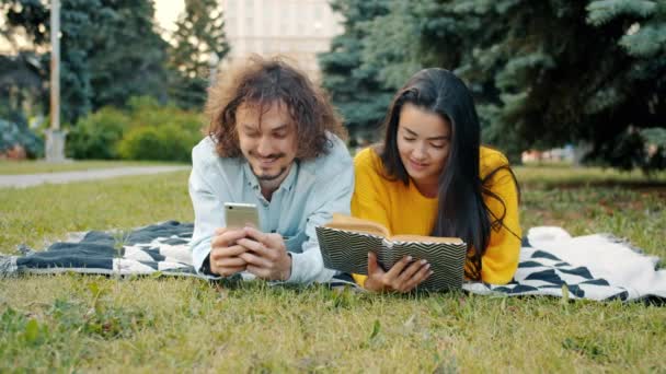 Hombre y mujer relajándose en el parque usando teléfono inteligente y lectura libro hablando — Vídeos de Stock