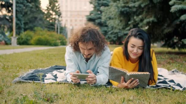 Ζευγάρι χαλαρωτικό σε εξωτερικούς χώρους στο πάρκο με το σκυλί παίζει το παιχνίδι smartphone και ανάγνωση — Αρχείο Βίντεο