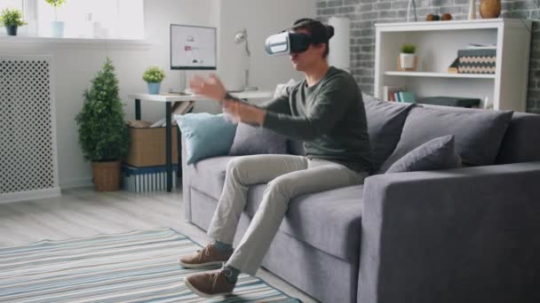 Χαρούμενος άντρας που κουνάει τα χέρια του καθισμένος στον καναπέ φορώντας γυαλιά εικονικής πραγματικότητας στο σπίτι — Αρχείο Βίντεο