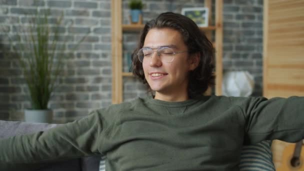 Улыбающийся молодой человек, вытянув руки, сидит на диване в квартире, расслабляясь дома — стоковое видео