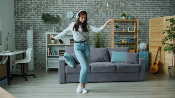 Hübsche junge Frau mit Kopfhörern tanzt in Wohnung und hat Spaß allein zu Hause — Stockvideo