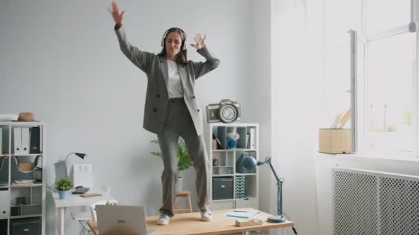 Vacker kontorsarbetare i kostym dansar på skrivbordet på kontoret bär hörlurar — Stockvideo
