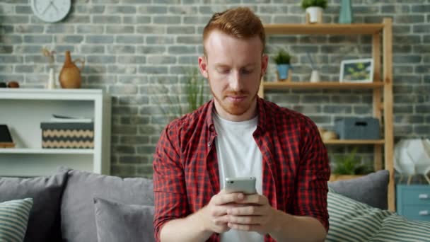 Vermelho barbudo cara mensagens de texto com smartphone sorrindo em apartamento aconchegante — Vídeo de Stock